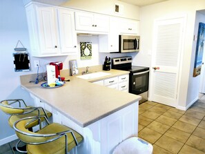 A kitchen with white cabinets, a microwave, a stove, and a sink. There are three yellow chairs and a trash can.