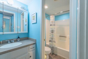 A bathroom with a blue wall, a white sink, a mirror, and a shower curtain with text. - Sunchase 206| Pool & Hot Tub | Steps to the Beach (Gulf Shores)