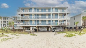 Un motel frente a la playa con varios balcones, una playa arenosa en primer plano y varios coches aparcados.