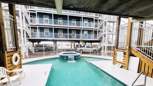 Zona de piscina con jacuzzi, sillones de descanso y vista a un edificio de varios pisos con balcones.