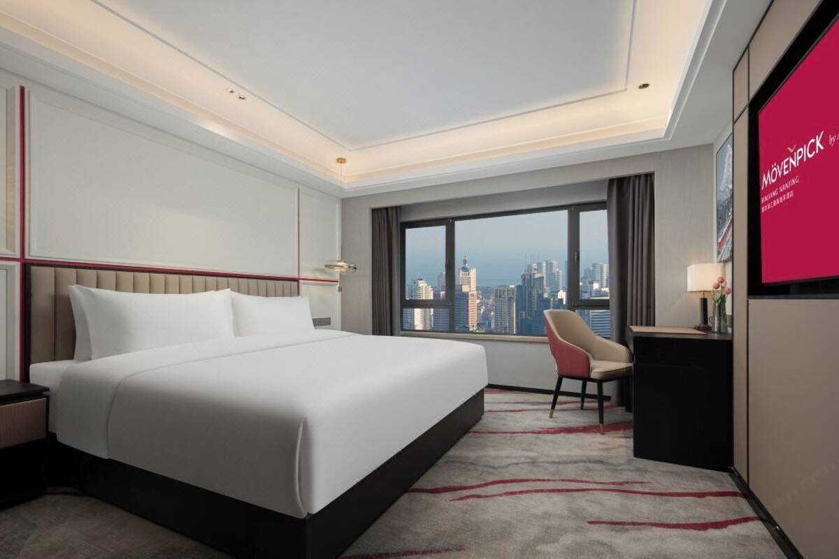 A modern hotel room with a large bed, a desk, a chair, and a city view.
