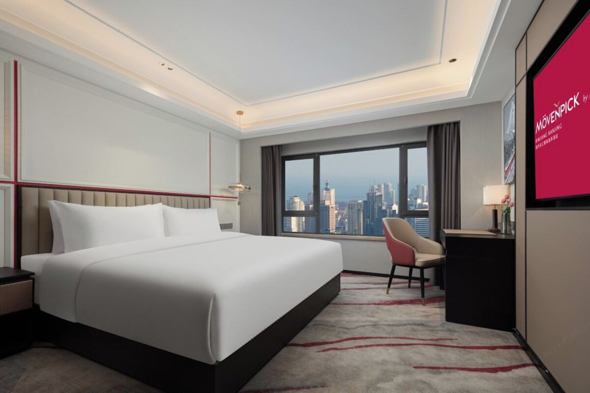 A modern hotel room with a large bed, a desk, a chair, and a city view.