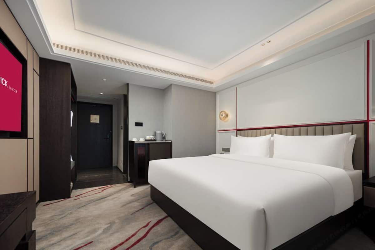 A modern hotel room with a large bed, a bedside table with a lamp, and a view of the hallway.