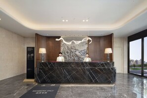 A modern hotel reception desk with two staff members, a marble counter, and a large window offering a city view. - Mövenpick By Accor Binjiang Nanjing (Nanjing)