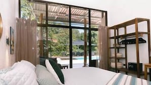 Ein Schlafzimmer mit Bett, Blick auf einen Pool und einem Holzregal mit Gegenständen.