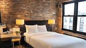 A modern bedroom with a brick wall, a large bed, two bedside lamps, a desk with a chair, and a view through a window. - Kosher House (Brooklyn)