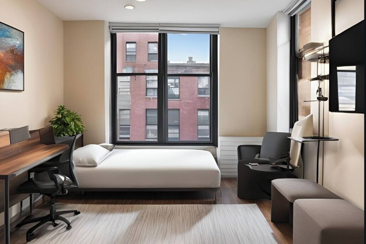A modern hotel room with a large window, a bed, a desk, a chair, and a small table.