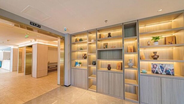 A modern hotel lobby with a central reception desk, multiple seating areas, and a large, well-lit shelving unit displaying various decorative items. - GreenTree Inn Express Hotel (Beijing)