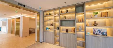 A hotel lobby with a reception desk, a staircase, and a wall with shelves displaying various items.