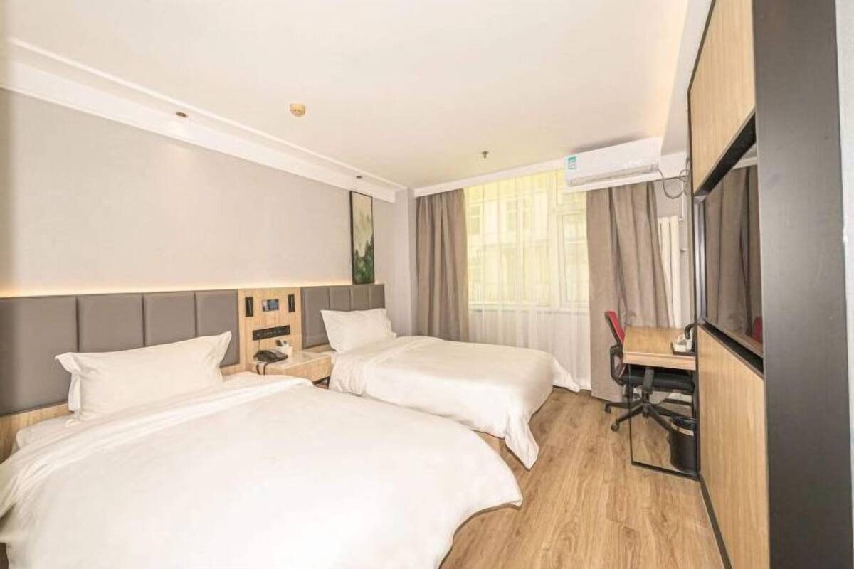 A hotel room with two beds, a desk, a chair, a TV, and an air conditioning unit.