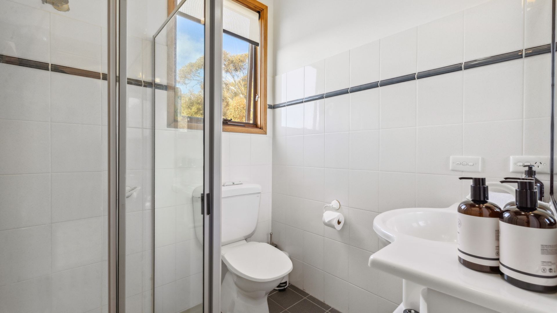 Ein Badezimmer mit weißen Fliesen, einer Duschkabine aus Glas, einer Toilette und einem Waschbecken mit zwei Seifenspendern.