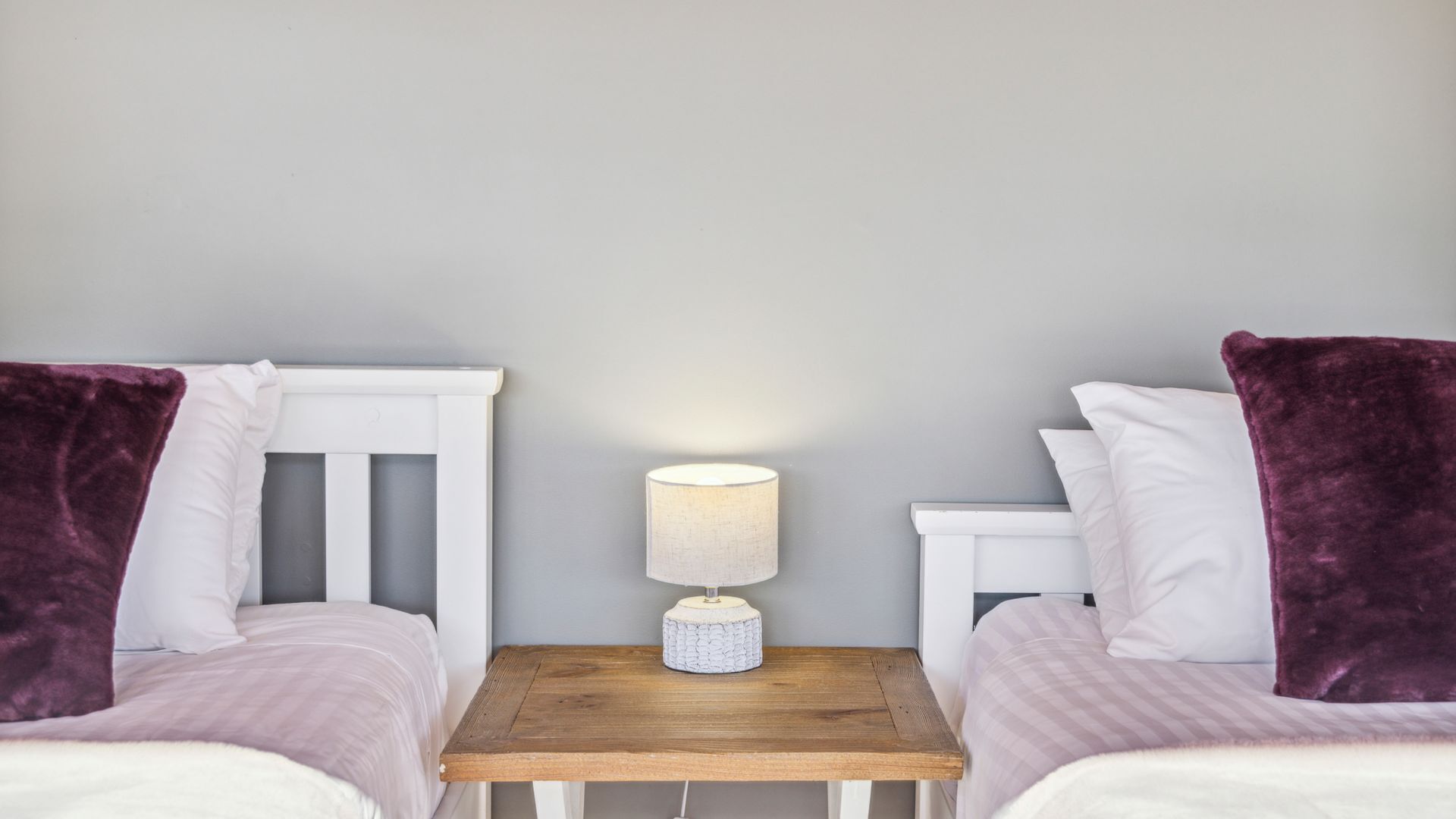 Een tweepersoonskamer met twee bedden, een nachtkastje met een lamp en een grijze muur.
