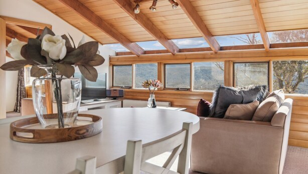 A cozy living room with a wooden ceiling, a round dining table, a sofa with cushions, and a large window offering a view of the outdoors. - Nowhere Apartment Crackenback (Crackenback)