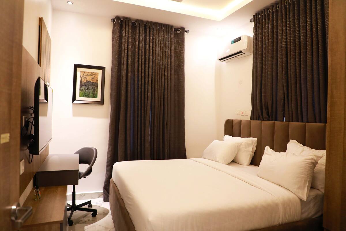 A hotel room with a bed, a desk, a chair, and an air conditioning unit.
