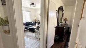 Een gang die leidt naar een woonkamer met een bank, een salontafel en een lamp. Aan de rechterkant een deur en links een plant.