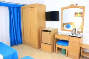 Ein Hotelzimmer mit Holzschrank, Schreibtisch mit Spiegel, Wandfernseher und einem Bett mit blauer Bettwäsche.