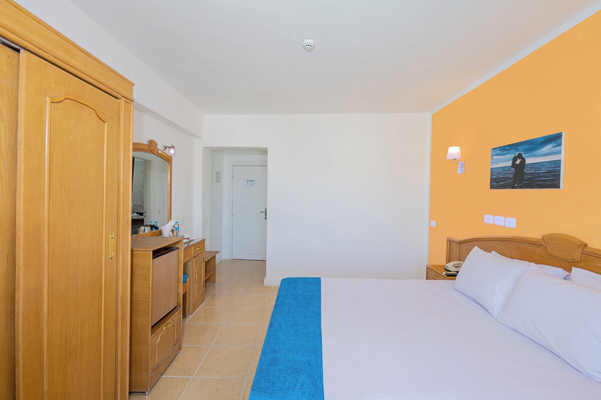 A hotel room with a bed, a wardrobe, a mirror, a desk with a lamp, and a picture on the wall.