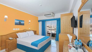 A hotel room with a bed, bedside table, and a desk with a mirror. There is a window with blue curtains, a view of buildings outside, and a ceiling fan. - Saint Maria Hotel Hurghada (Hurghada)