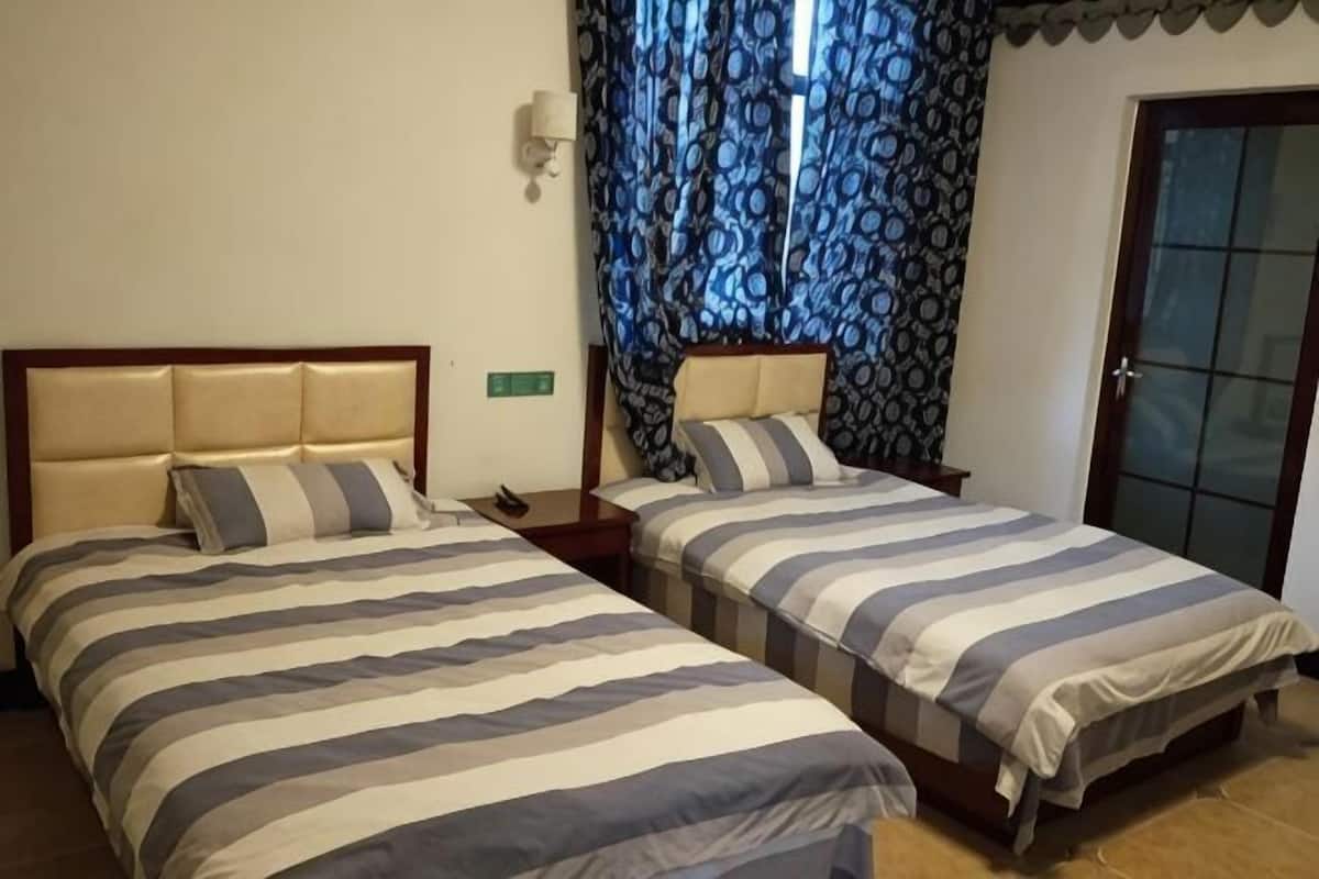 Two single beds with striped bedding, a nightstand, a wall-mounted lamp, and a patterned curtain.