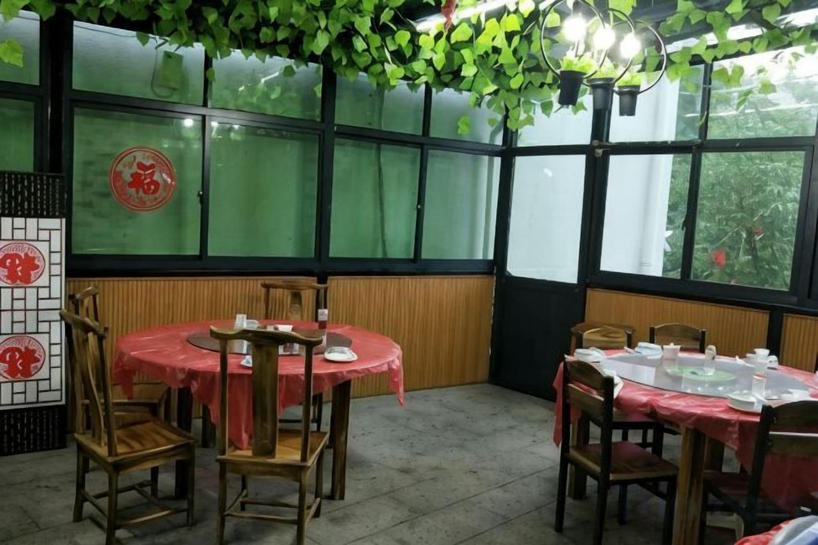 Un restaurante con mesas y sillas redondas, una pared verde con un emblema rojo y grandes ventanales.