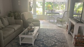 A living room with a sofa, a coffee table, a chair, a television, and a view of the outdoors through a glass door. - 1st Floor Charming 2-bedroom condo in Bonita Springs with Community Pool (Bonita Springs)