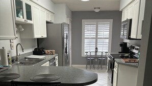 A kitchen with white cabinets, a granite countertop, a bar area with stools, and a window with blinds. - 1st Floor Charming 2-bedroom condo in Bonita Springs with Community Pool (Bonita Springs)