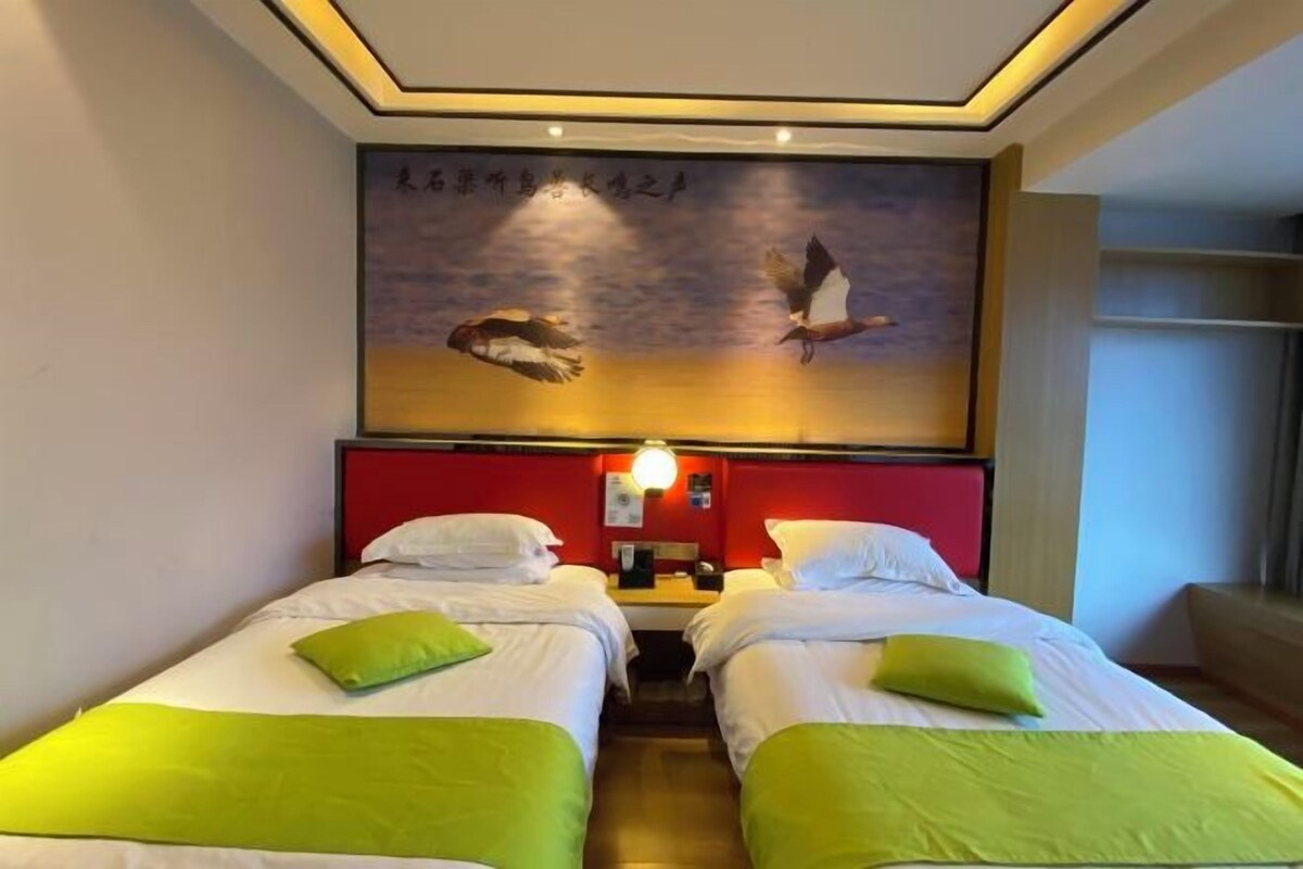 A hotel room with two beds, a red headboard, a wall-mounted artwork, and a bedside table with a lamp.