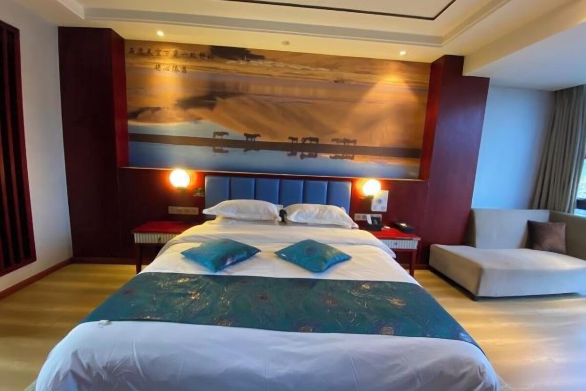 A hotel room with a large bed, a headboard with a mural, bedside lamps, and a seating area with a chair and a small table.