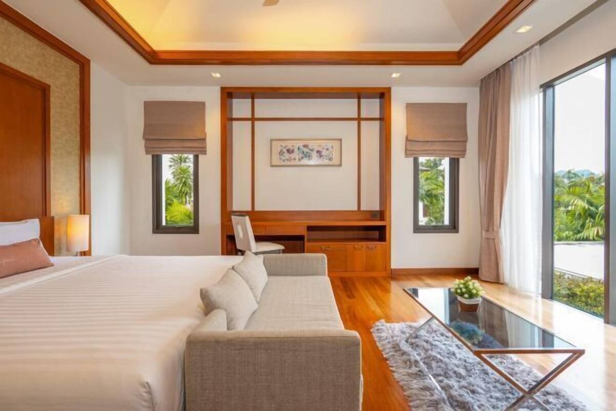 A modern hotel room with a large bed, a sofa, a desk, and a view of greenery.
