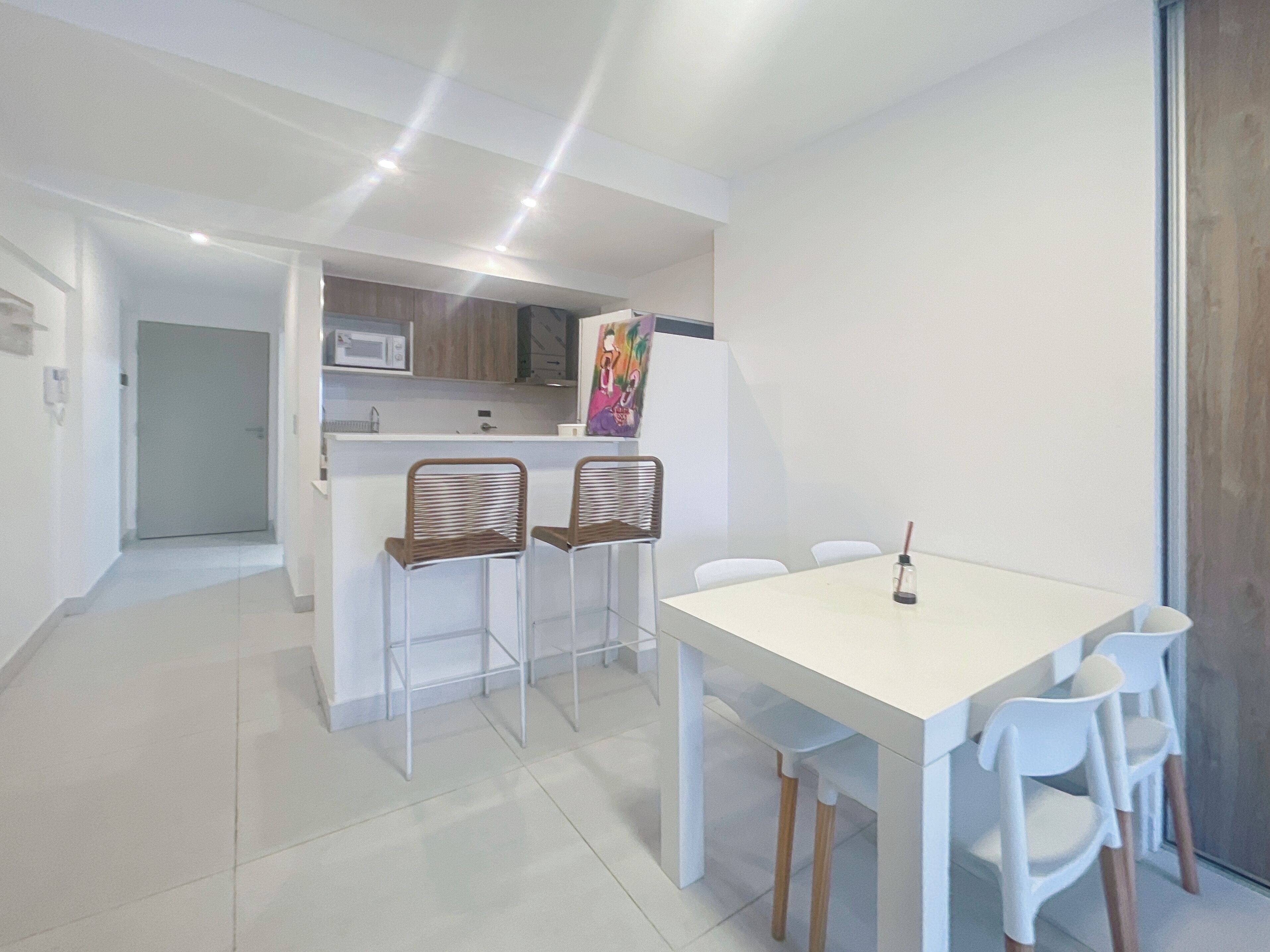 Una cocina moderna con armarios blancos, microondas y refrigerador. Hay un comedor con una mesa blanca y cuatro sillas.