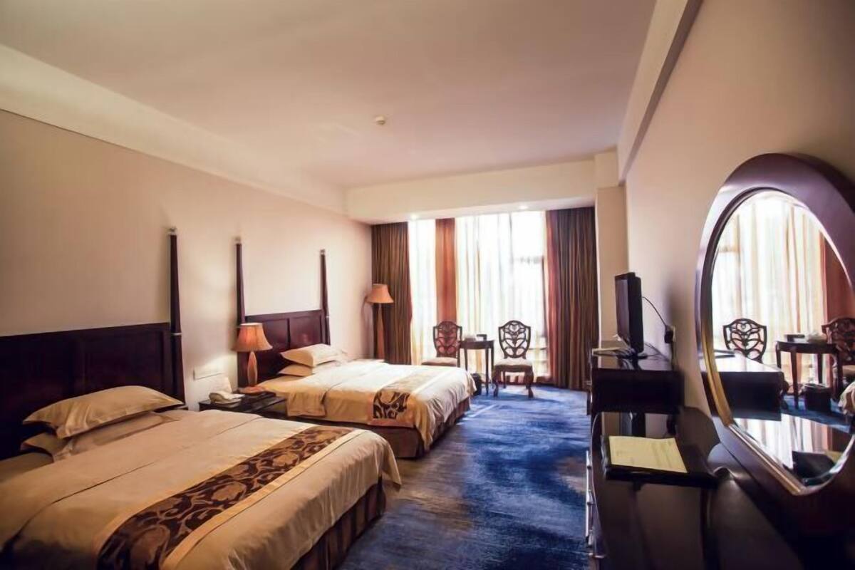 A hotel room with two beds, a desk, a chair, a mirror, and a large window with curtains.