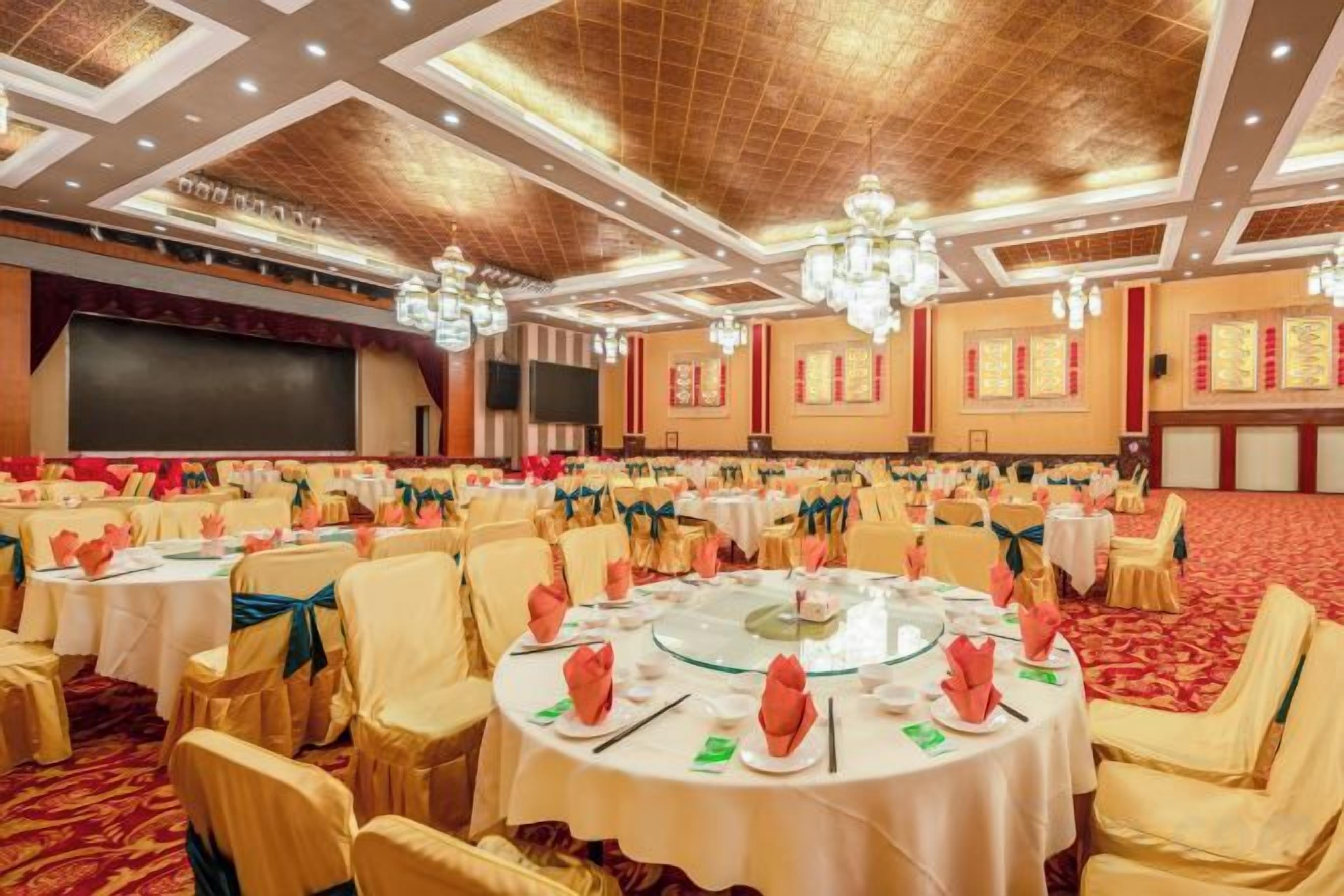 Un salón de banquetes con mesas redondas dispuestas para comer, sillas y un escenario con una pantalla.