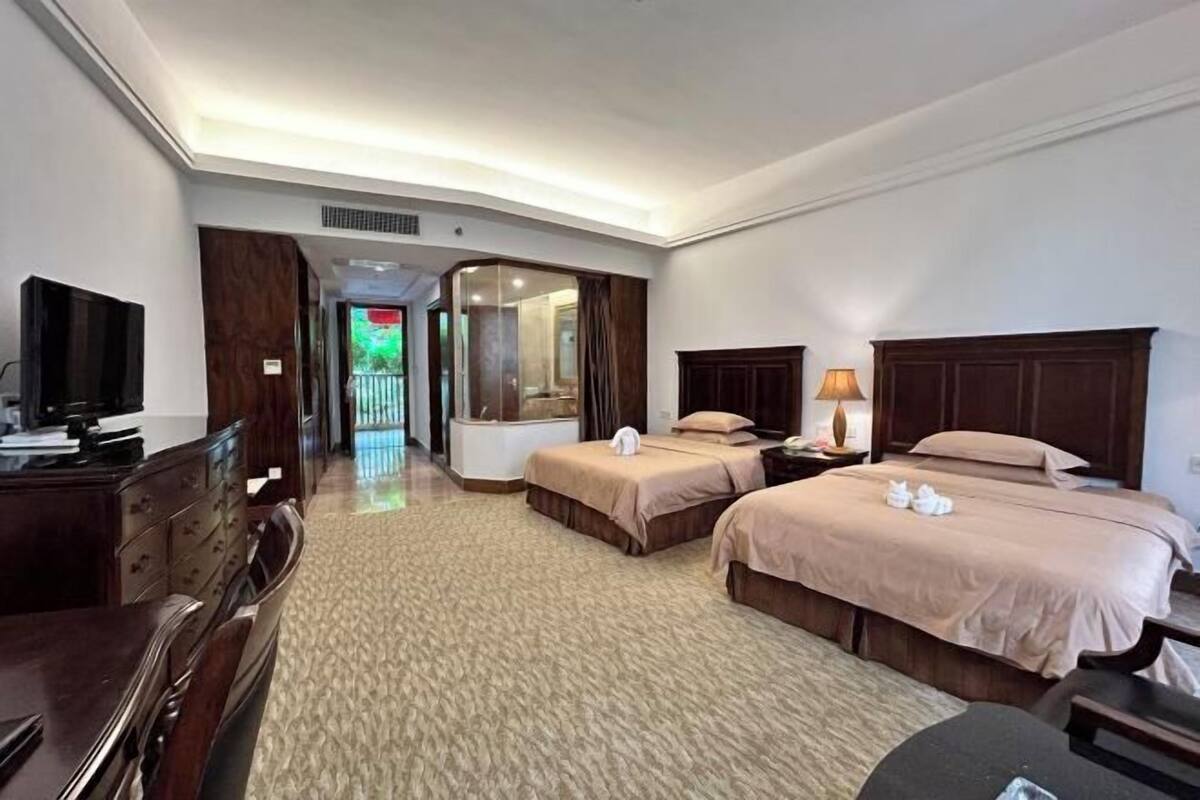 A hotel room with two beds, a desk with a chair, a TV, and a bathroom visible through a glass door.