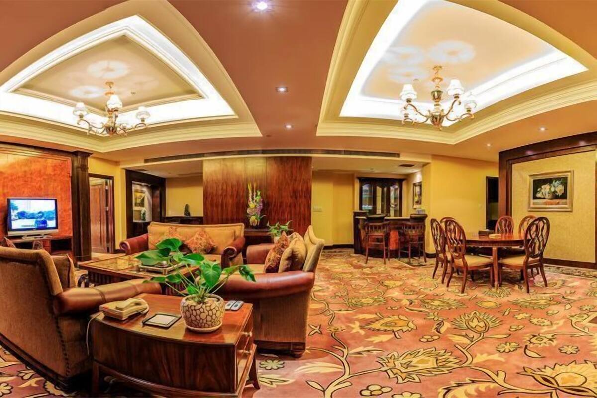 A spacious lobby with a high ceiling, a central seating area with sofas and a coffee table, a dining area with a wooden table and chairs, and a television on the left.