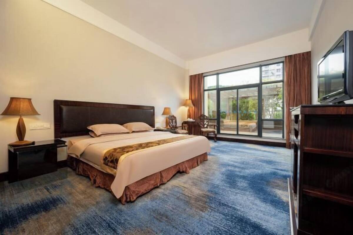 A hotel room with a large bed, a TV, a desk, and a view of the outdoors through a large window.