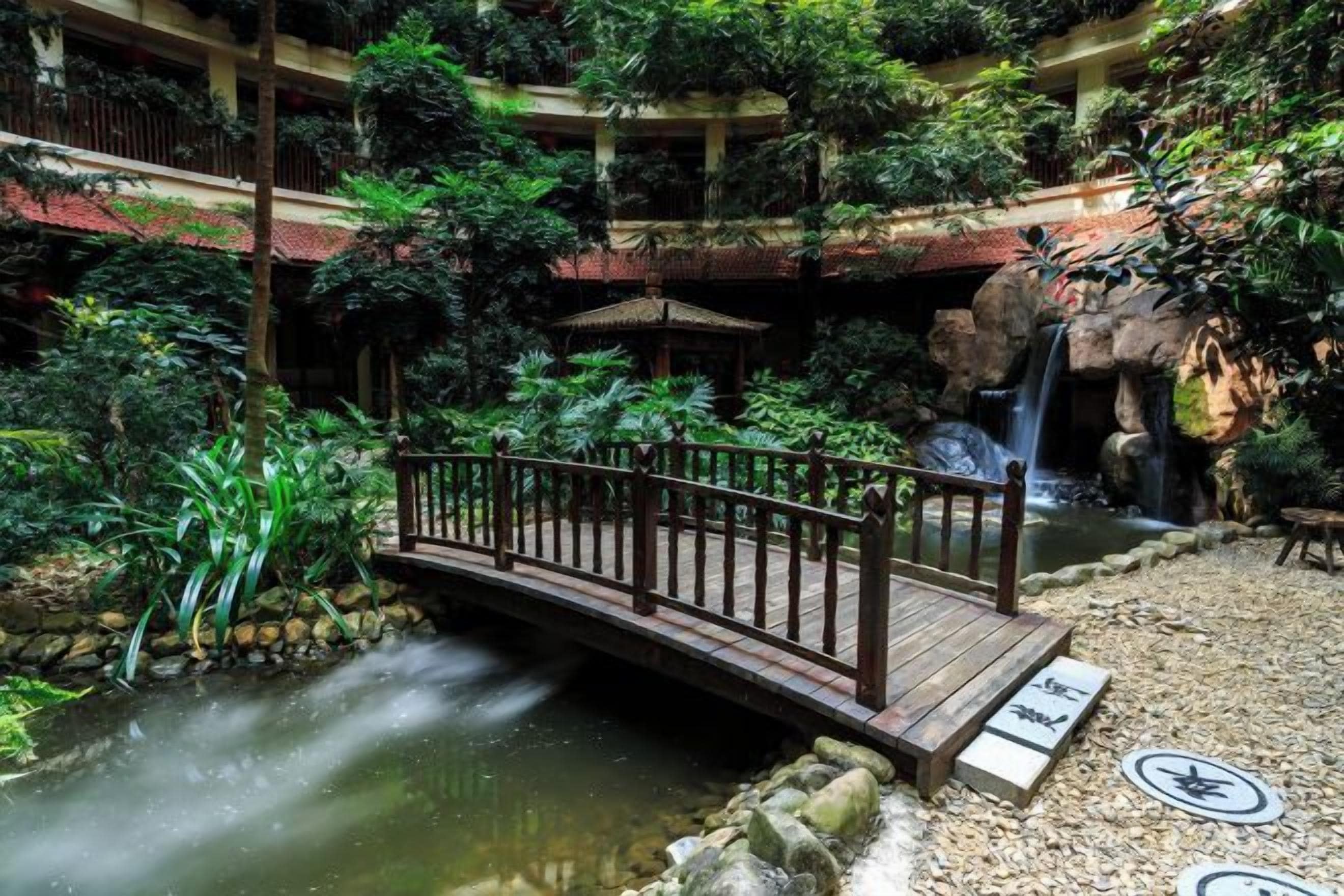 Ein mehrstöckiger Innenhofgarten mit einer Holzbrücke über einem Bach, umgeben von üppigem Grün und einem Wasserfall.