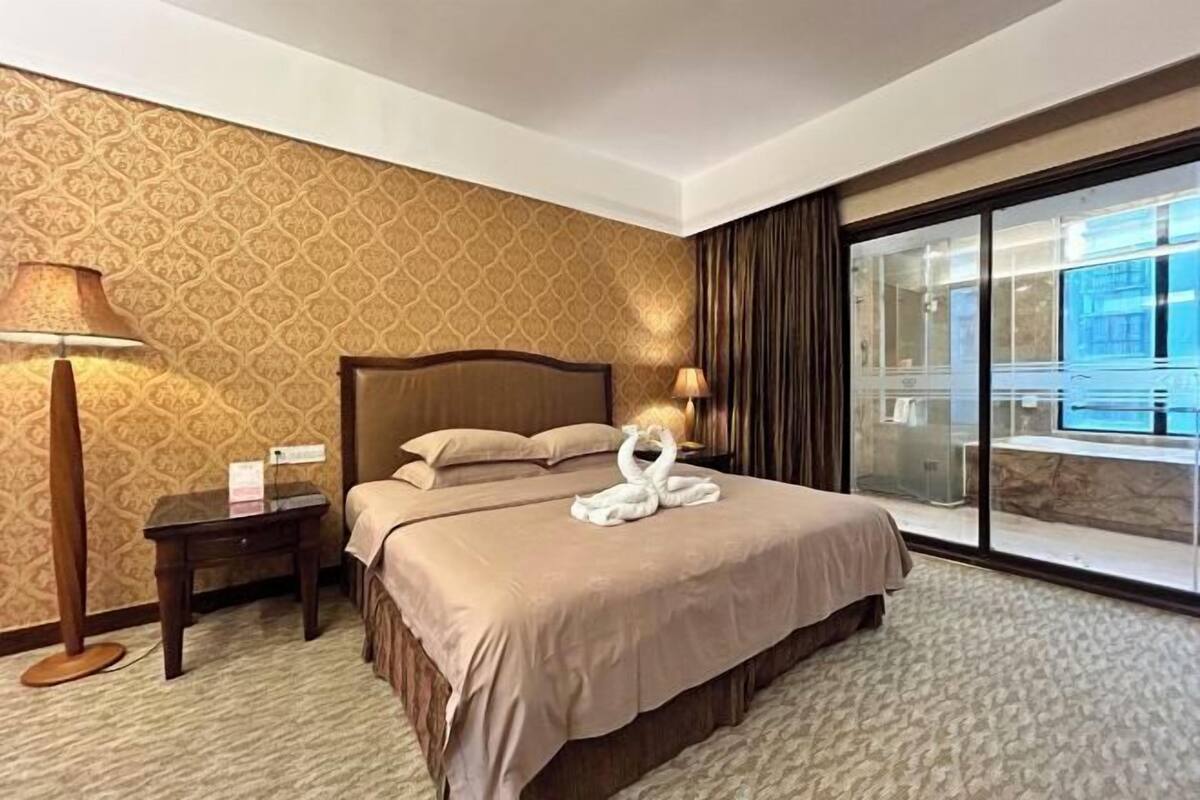 A hotel room with a large bed, a nightstand, a lamp, and a view of a pool through a glass door.