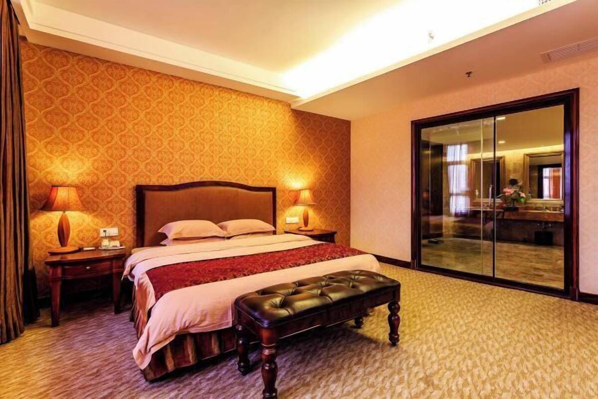 A hotel room with a large bed, two bedside lamps, a wooden bench, a patterned carpet, and a large mirror with a sliding door.
