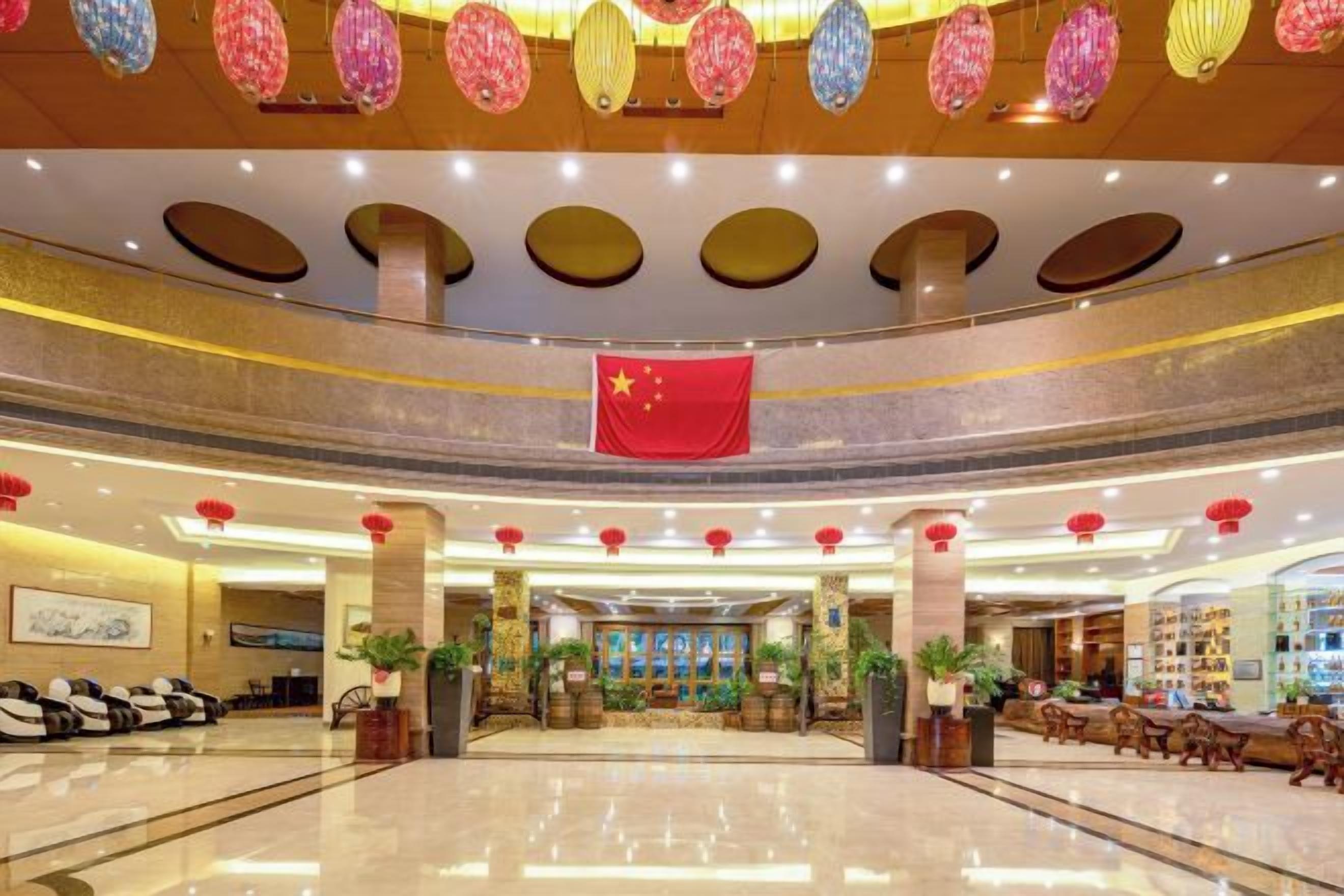 Un vestíbulo de hotel con mostrador de recepción central, zona de estar y elementos decorativos como faroles colgantes y una bandera.