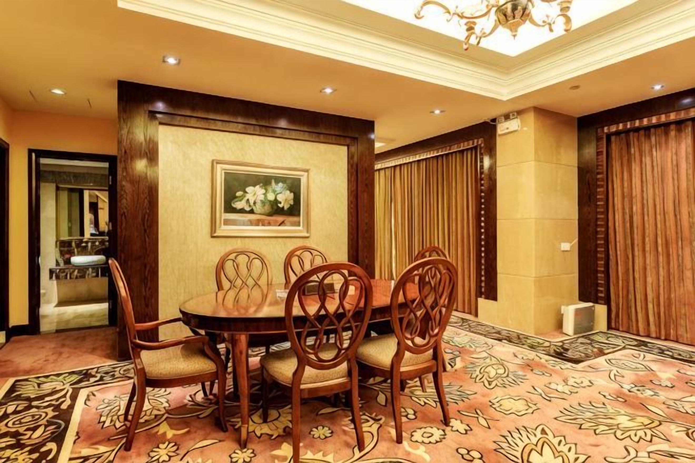 A dining area with a round table and six chairs, a chandelier, a framed floral painting, and a patterned carpet.