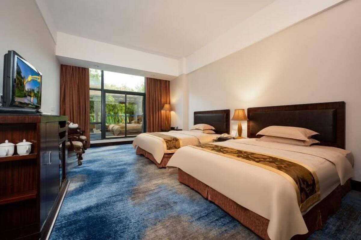 A hotel room with two beds, a television, a desk, and a view of greenery outside.