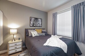 A bedroom with a bed, bedside table, lamp, clock, and a window with curtains. - Waterwalk Extended Stay Kansas City - Overland Pk (Overland Park)