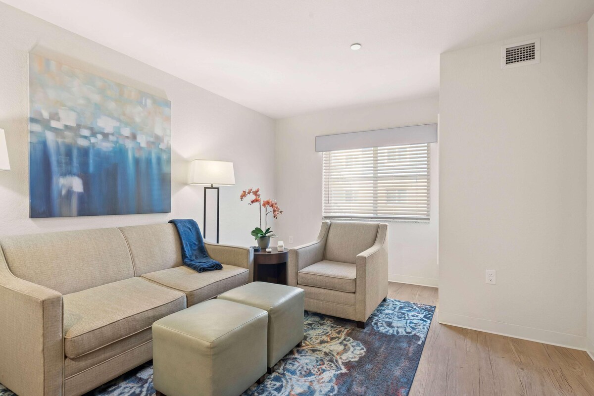Een woonkamer met een beige bank, een bijpassende fauteuil, een klein bijzettafeltje met een lamp en een groot abstract schilderij aan de muur.