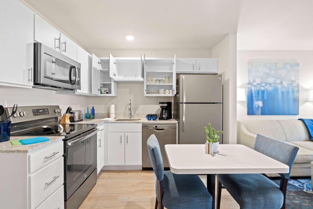 Una cucina moderna con elettrodomestici integrati, una zona pranzo con tavolo e sedie e un soggiorno con divano e un quadro appeso al muro.