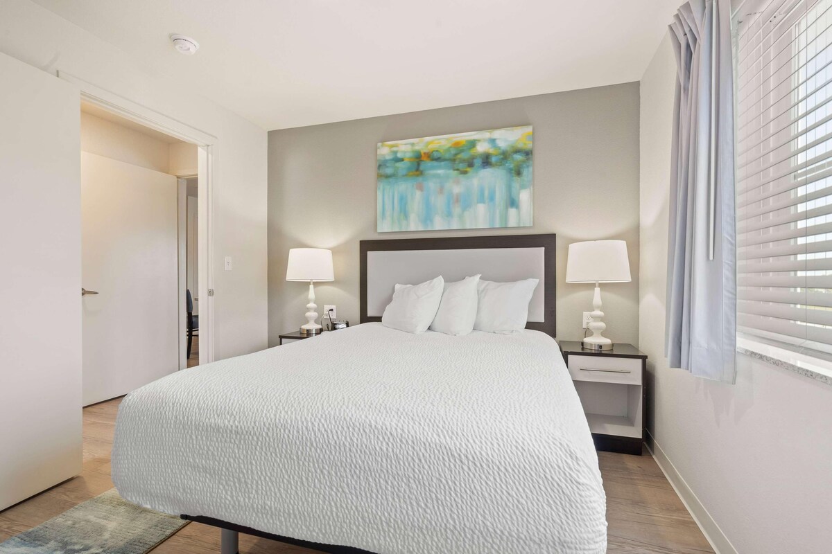 Une chambre d’hôtel bien rangée, avec un grand lit, deux tables de chevet avec des lampes, un tableau au mur et une fenêtre avec des stores.