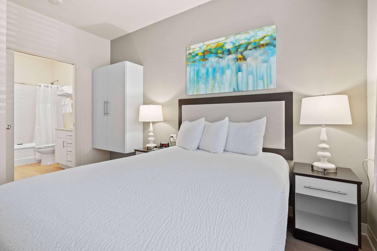 A hotel room with a large bed, two bedside tables with lamps, a wardrobe, and a bathroom visible through an open door.
