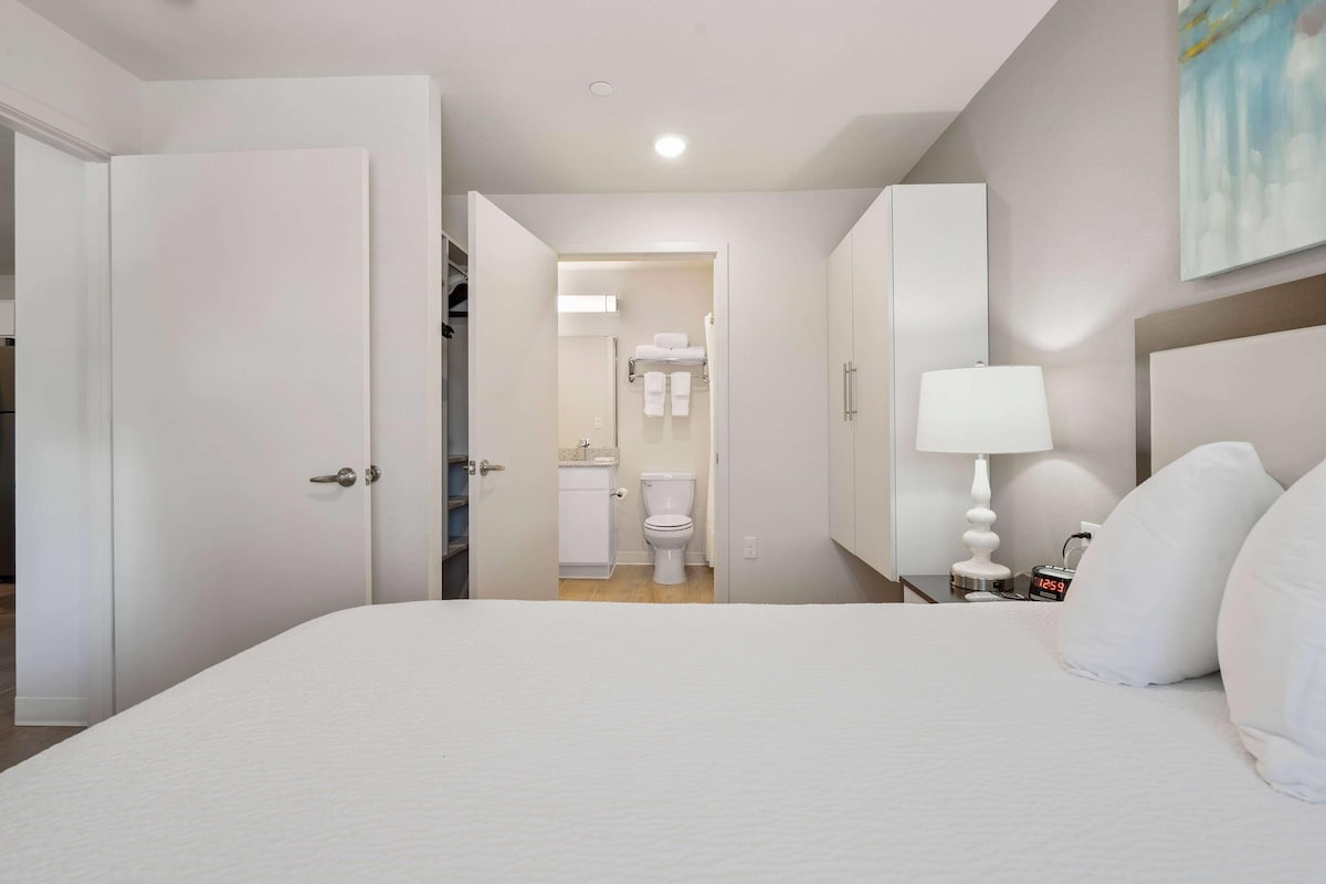 Une chambre d’hôtel avec un grand lit, une table de chevet avec une lampe, une commode et vue sur la salle de bain.