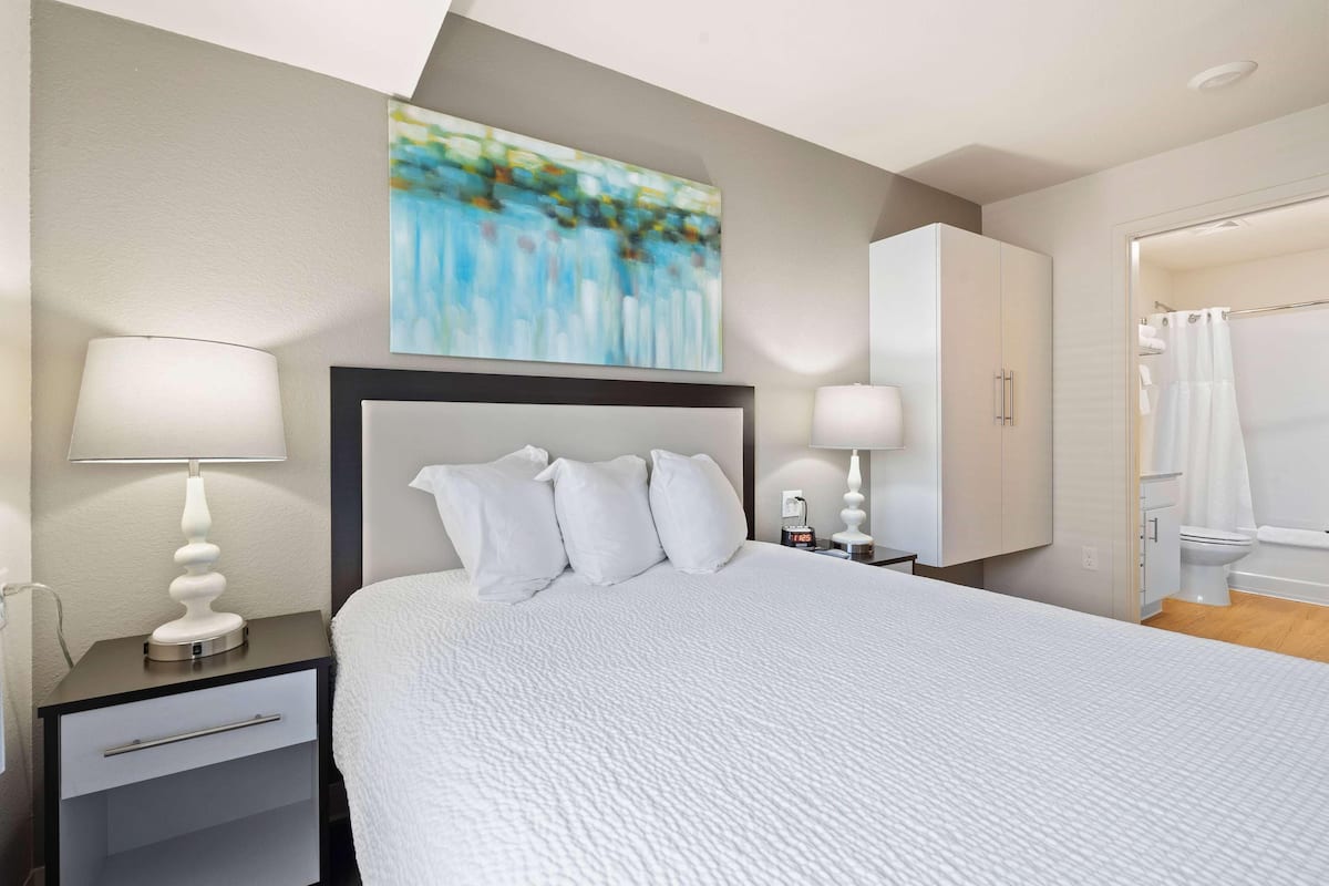 A neatly arranged bedroom with a bed, bedside lamps, a nightstand, a wardrobe, and a bathroom visible through an open door.