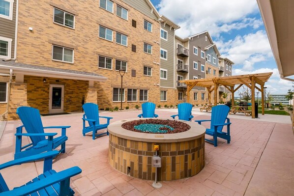 A courtyard with a fire pit surrounded by blue Adirondack chairs, a wooden pergola, and a brick building with multiple windows and balconies. - Waterwalk Extended Stay Kansas City - Overland Pk (Overland Park)