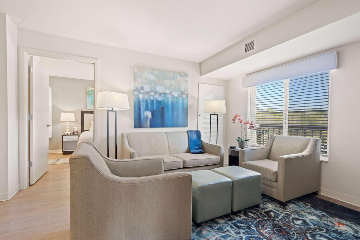 A modern living room with a sofa, armchairs, and a coffee table. There is a painting on the wall, a lamp, and a window with blinds.